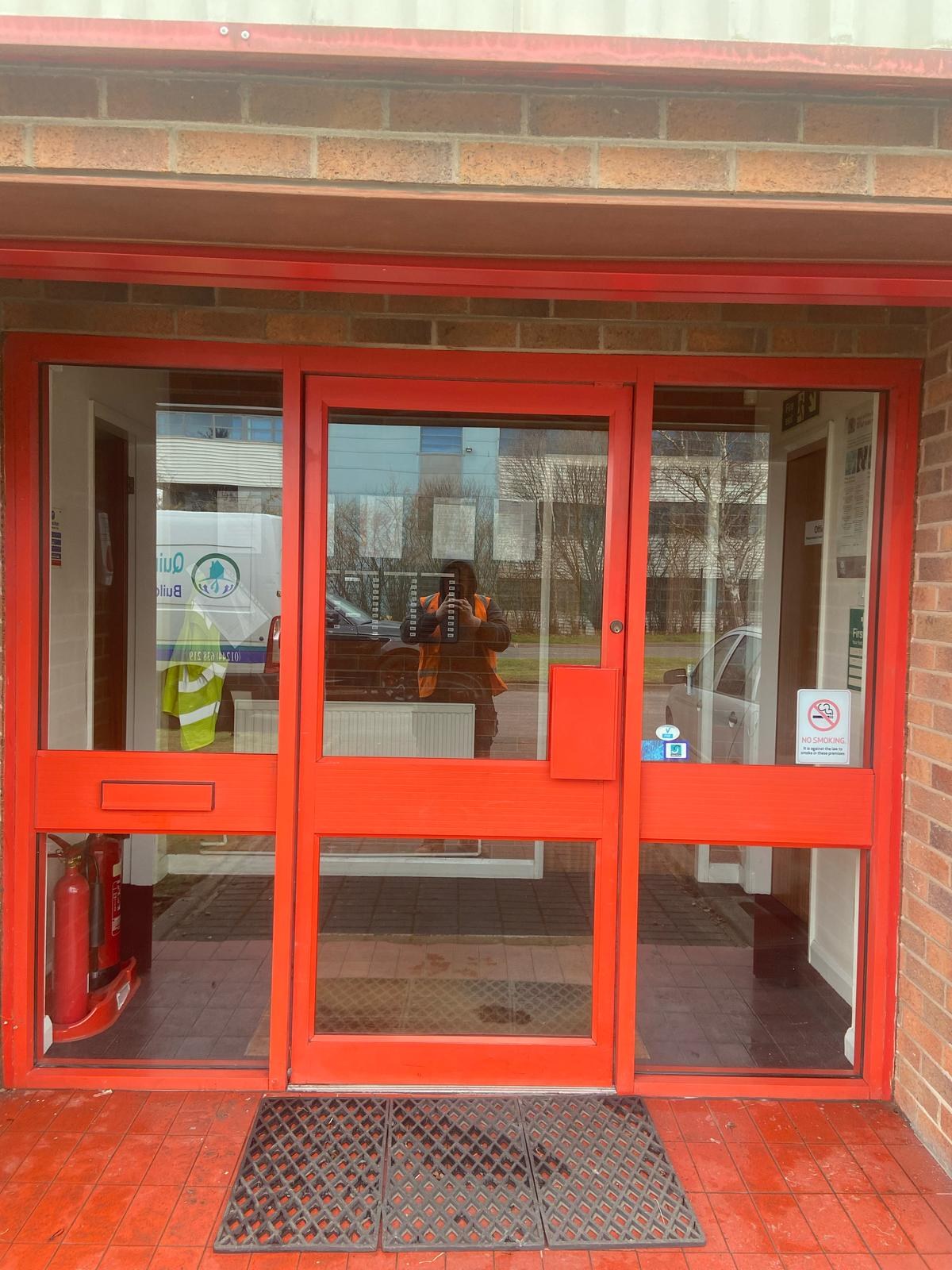 Red-framed glazed entrance door closed, with a Quirks van and operative reflected in the glass