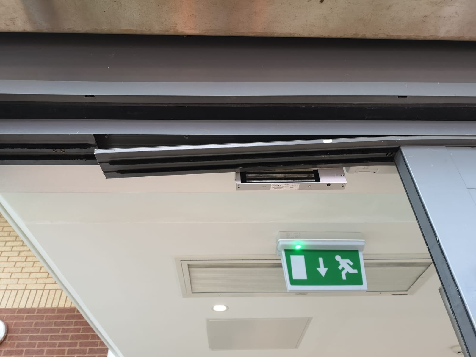 Head of a metal-framed entrance door from below, with a trim section hanging loose beside a magnetic lock