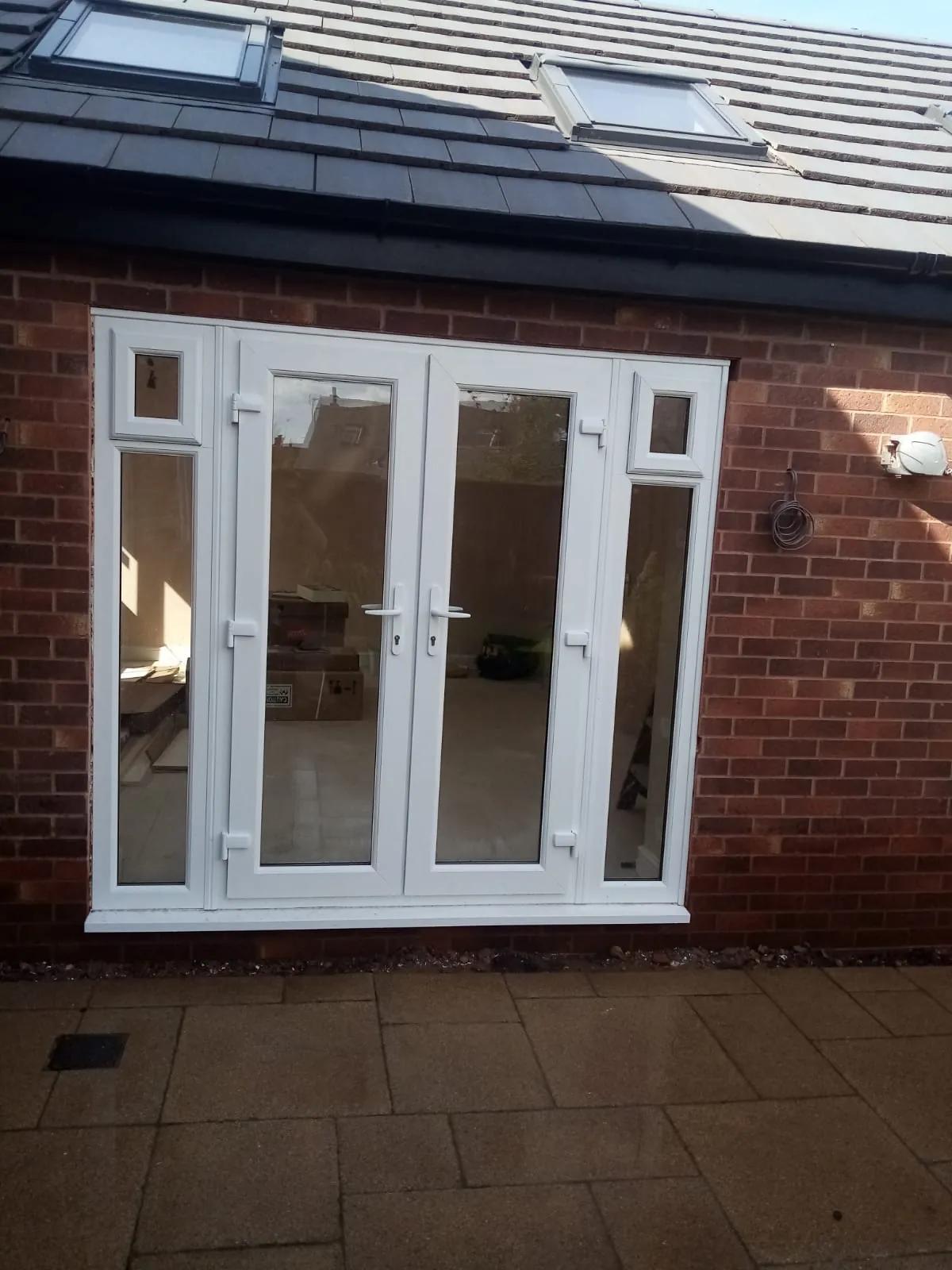 White uPVC French doors with side panels set in a red brick wall beneath a pitched roof with roof windows