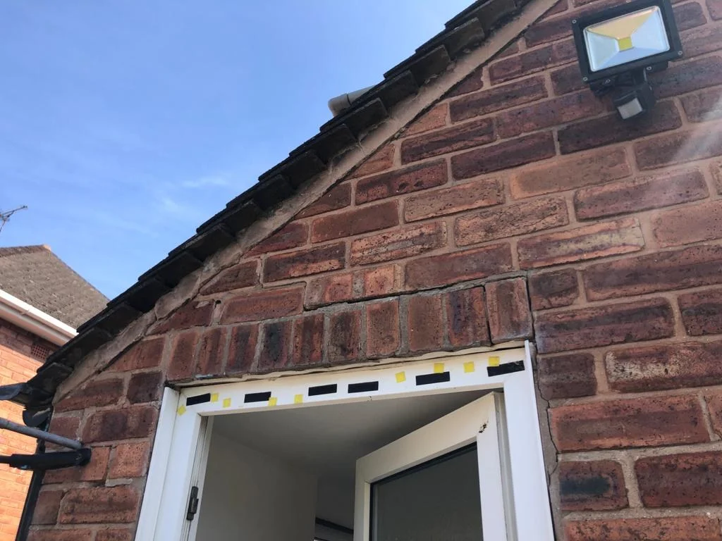 Course of bricks on end above a white door frame, dropped out of line with cracked mortar joints, under the edge of a tiled roof