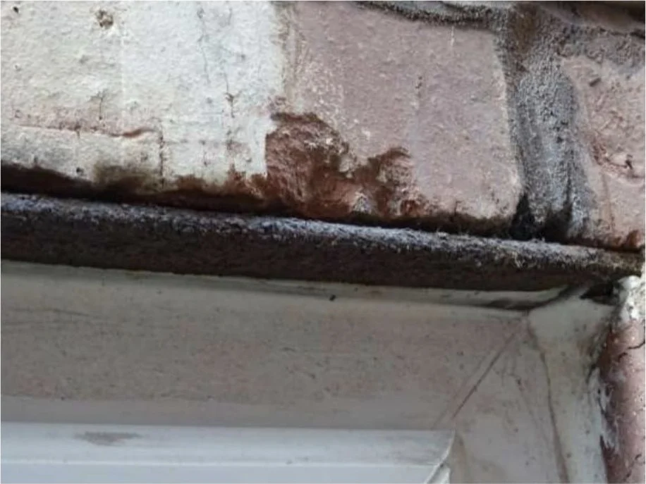 Heavily corroded steel lintel above a window head, with rust staining and salt deposits on the brickwork above it