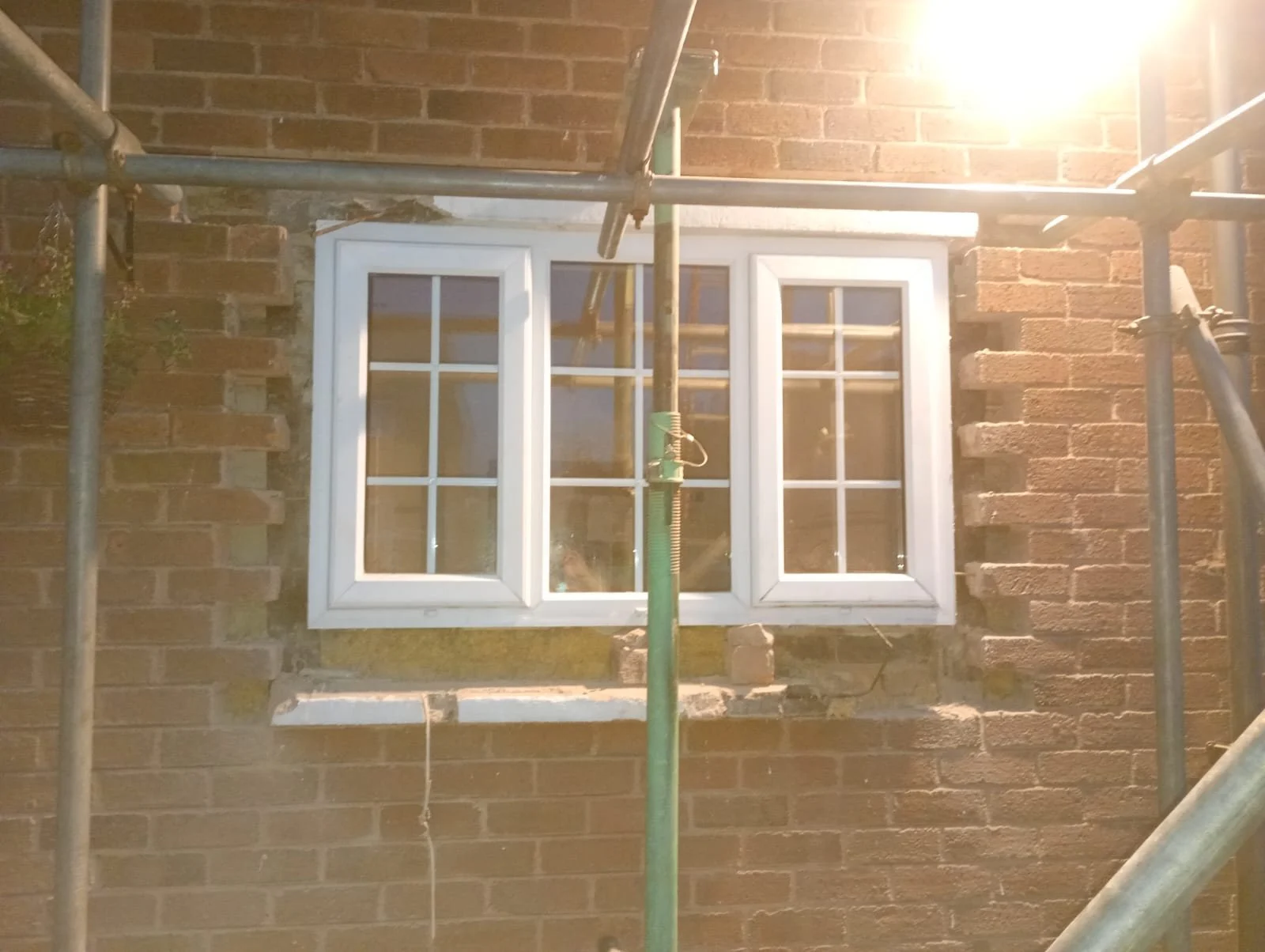 White three-pane window seen through scaffolding, with the surround removed, brickwork toothed back either side and a prop in front