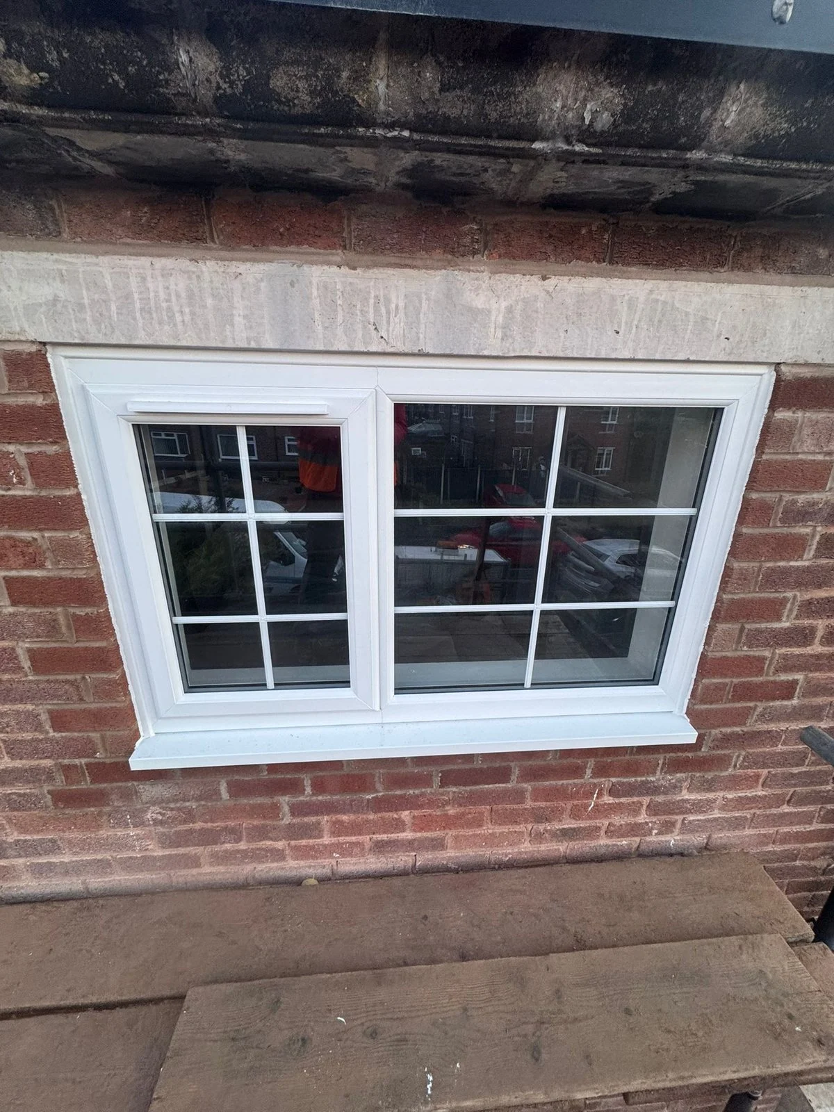 New white uPVC window under a concrete lintel in a red brick wall, seen from a scaffold