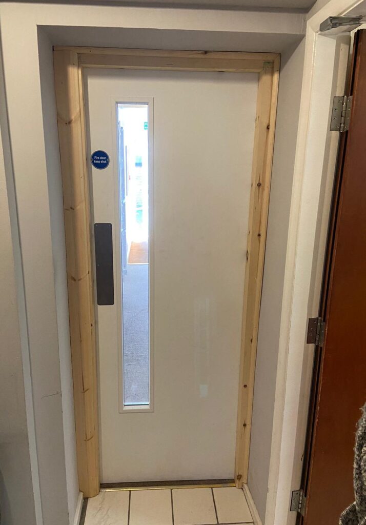 White FD30 corridor fire door with a narrow vision panel, push plate and fire door keep shut sign, in an undecorated timber frame
