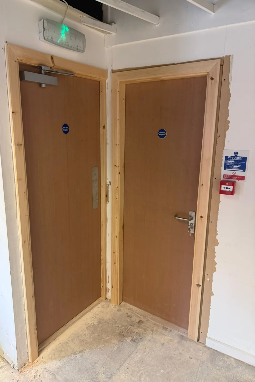 Two plywood-faced fire doors in new softwood frames at the corner of a corridor, one with an overhead closer and push plate, beneath an emergency light Two plywood-faced fire doors in new softwood frames at the corner of a corridor, one with an overhead closer and push plate, beneath an emergency light