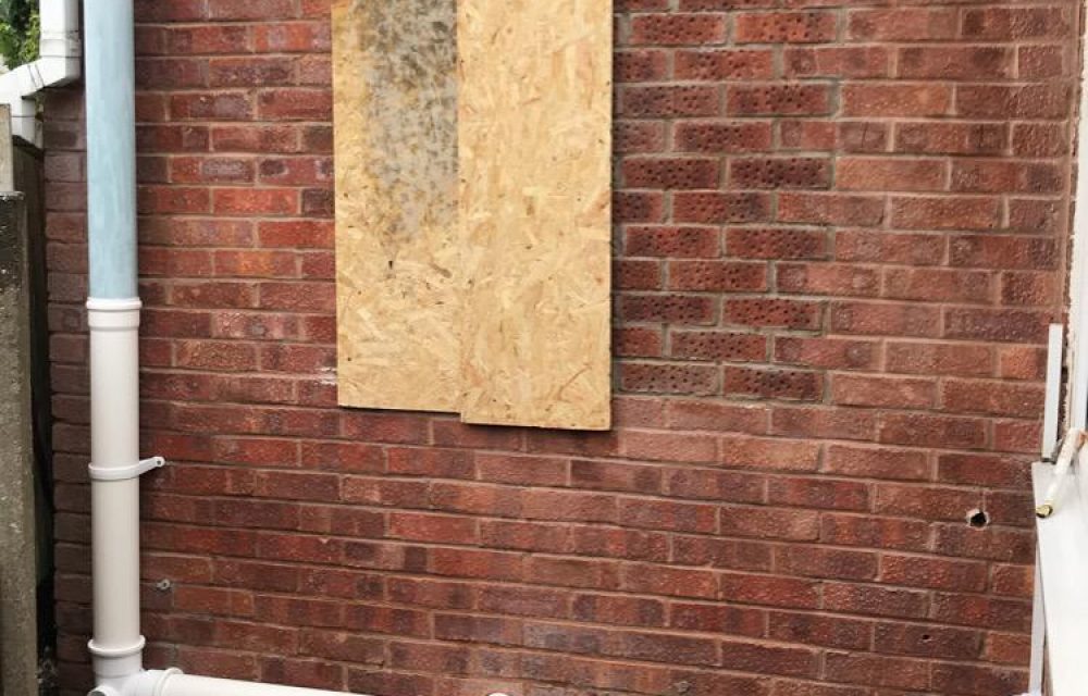 Small window on a side elevation boarded up with OSB beside the downpipes